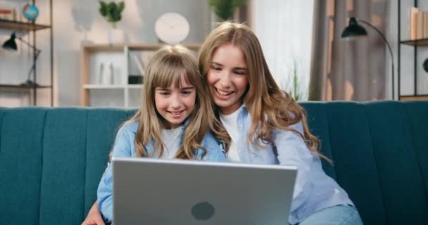 Close up portret piękny uśmiech szczęśliwy beztroski dwie siostry, które siedzą na kanapie w domu i machając rękami do kamery internetowej, aby powiedzieć hallo do rozmówcy podczas czatu wideo na laptopie — Wideo stockowe
