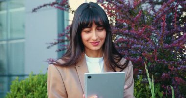 Modern bir ofis binasının yakınında elinde tabletle gülümseyen genç bir kadının yakın çekimi, iş ortamında bir şehir caddesinde duruyor. Yavaş çekim