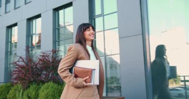 Modern cam ofis binasının yanında güvenle yürüyen genç bir beyaz kadının yandan görünüşü elinde dizüstü bilgisayar ve defter taşırken gülümsüyor. Neşeli ve işe gitmeye kararlı. Yavaş çekim