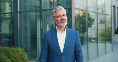 Şehrin dışında duran, kolları bağlı kameraya güvenle gülümseyen, arka planda modern cam ofis binası olan kıdemli sakallı iş adamının portresi. Yavaş çekim