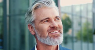 Dışarıda duran gri saçlı, arka planda modern cam ofis binası olan kameraya içten bir şekilde gülümseyen yaşlı sakallı bir adamın portre yüzü. Şehir işi ya da yaşam tarzı konsepti... Yavaş çekim
