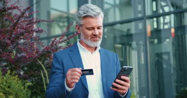 Yetişkin bir adam cam ofis binasının yanında otururken internet bankacılığı için akıllı telefon ve kredi kartı kullanıyor. Fintech, dijital işlemler ve mobil para yönetimi kavramı. Yavaş çekim