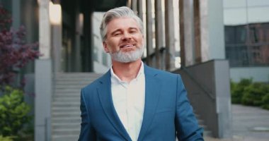 Şehir caddesinde yürüyen kıdemli işadamı, arka planda modern cam ofis binasıyla kameraya gülümsüyor. Liderlik, deneyim, güven ve profesyonel başarı kavramı. Yavaş çekim