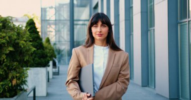 Uzun siyah saçlı profesyonel bir kadın dizüstü bilgisayar tutuyor ve son moda bir çalışma alanında duruyor. Açık havada, dostça bir gülümsemeyle kameraya bakıyor.