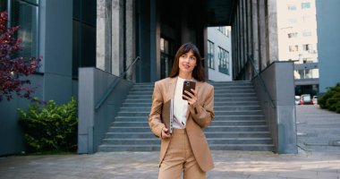 Şehirde bir iş gününden sonra modern bir ofis binasının önünde dikilirken resmi kıyafetli gülümseyen profesyonel bir kadın akıllı telefon kullanıyor.