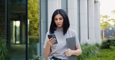 Siyah saçlı, dizüstü bilgisayarı olan ve telefon kullanan çekici bir genç kadın. Açık havada.