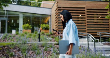 Gündelik kıyafetli beyaz bir iş kadını, şehir parkındaki modern bir binanın önünde yürürken akıllı telefon ve dizüstü bilgisayar kullanıyor. İş ve insan kavramları