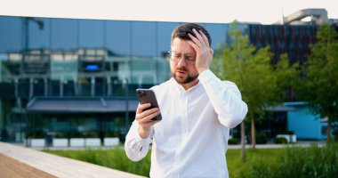 Beyaz tişörtlü, bekar, cep telefonunu kontrol eden, modern cam bina arka planında, kapalı alanda yürüyen bir adam.