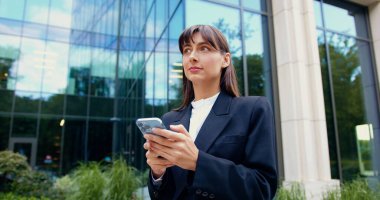 Siyah ceketli gülümseyen esmer kadın akıllı telefon kullanıyor ve açık havada güneşli bir yaz gününde modern cam bir binanın önünde yürürken mesaj yazıyor..