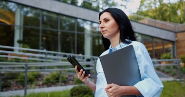 Kentli bir parkta elinde dizüstü bilgisayarla gezen ve arka planda modern şehir mimarisi olan akıllı telefon kullanan kendine güvenen genç bir kadının yan görüntüsü. Hareketlilik ve dijital yaşam tarzı kavramı