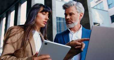İki iş adamının, resmi giyinmiş bir kadın ve bir erkek, modern bir ofis binasının önünde dikilip dijital tablet ve dizüstü bilgisayar kullanarak işle ilgili konuları tartışıyorlar.