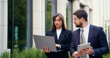 Odaklanmış bir iş kadını, elinde tablet bulunan başarılı erkek arkadaşıyla birlikte bir şehir bölgesinde yürürken dizüstü bilgisayarını işaret ediyor. Birlikte yeni bir projeyi tartışıyorlar.
