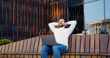 Beyaz gömlekli bir iş adamı bankta oturuyor ve modern cam binanın yanında dizüstü bilgisayarla çalışıyor. Odaklanmış genç adam uzaktan çalışıyor.