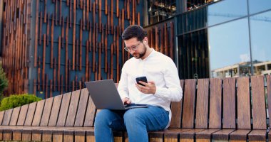 Modern ofis çalışanı, ofis binasının etrafında otururken dizüstü bilgisayar ve telefon kullanıyor. Odaklanmış iş adamı dışarıda uzaktan çalışır..