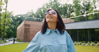 Tatmin olmuş genç bir kadın yeşil bir parkta sakin bir yürüyüş yaparken temiz havanın tadını çıkarıyor. Rahatlamış ve mutlu görünüyor, güneşli bir günde doğayla iç içe.