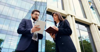 İş adamı ve resmi takım elbiseli iş kadını modern ofisin yakınlarındaki açık havada işbirliği yapıyor, program planlama, takım çalışması, beyin fırtınası ve rapor tartışmaları için tablet ve defter kullanıyorlar.