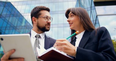 İki meşgul ve kendine güvenen iş adamının bir ofis binasının yanında durup tablet ve el yazısıyla yazılmış notlar kullanırken acil iş meselelerini tartışması.