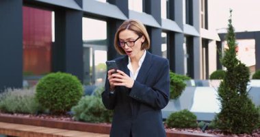 İş kadını dışarıda durmuş akıllı telefondan iyi haberlerle e-posta okuyor. Mutlu kadın zaferi kutluyor, piyango zaferi, açık havada açık havada hediye veriyor. Yavaş çekim.