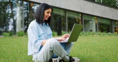 Yeşil şehir parkında dizüstü bilgisayarla çalışan genç bir kadın yemek molasında dijital yaşam özgürlüğünün tadını çıkarıyor..