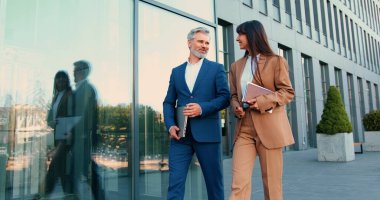 Resmi iş kıyafetleri giymiş yetişkin bir erkek ve genç bir kadın, dışarıda iş konuşmaları yaparken birlikte ofise doğru yürüyorlar.