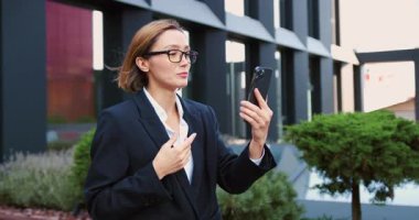Gözlüklü bir iş kadını şehir iş yerindeki akıllı telefondan el sallıyor. Müşteri ya da iş ortağıyla çevrimiçi görüşme yapan bir girişimci. Yavaş çekim.