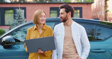 İki gülümseyen insanın portresi, kadın ve erkek, dizüstü bilgisayar tutuyorlar ve dışarıda elektrikli bir arabanın önünde poz veriyorlar. Modern insanlar, teknoloji ve çevre dostu yaşam tarzı kavramı. Yavaş çekim.