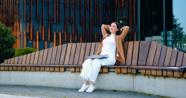 Kafkasyalı bir kız, sıradan kıyafetlerle bankta oturuyor, kolları başının arkasında, gülümsüyor ve laptopuyla dışarıda, kucağında dinleniyor.
