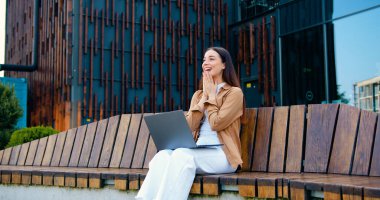 Gülümseyen kız elleri yüzü üzerinde bankta oturuyor, bilgisayarda çalışırken mutlu hissediyor, iş ve açık havada başarıya odaklanıyor.