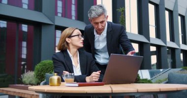 Profesyonel erkek kadın, açık kafede oturup dizüstü bilgisayar tartışması yapıyor. Bilgisayarlı çekici iş kadını iş adamını dinliyor. Hem gülümsüyor hem de güneşli bir şehirde etkileşime geçiyor. Yavaş çekim