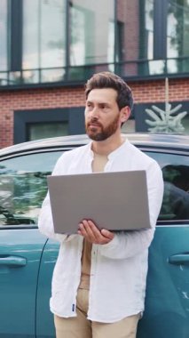 Şehir merkezindeki uzaktan kumandayla elektrikli arabası şarj olurken modern iş adamı dizüstü bilgisayarda çalışıyor. Sürdürülebilir hareketlilik ve dijital yaşam tarzının sembolü