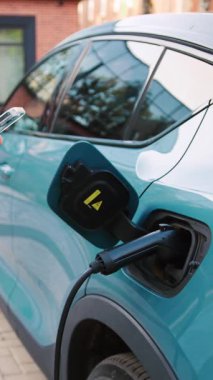 Şehir binalarıyla çevrili bir caddedeki taşınabilir istasyonda elektrikli arabasının yanında duran bir adam akıllı telefon uygulaması kullanıyor. Yavaş çekim