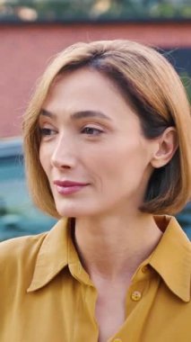 Açık havada duran kendine güvenen genç bir sarışının yakın çekimi. Profesyonel ve yaklaşılabilir bir ifadeyle kameraya bakıyor. Günlük kıyafetler giyiyor. Yavaş çekim