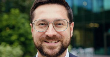 Gözlüklerini ayarlayan, arka planı bulanık, açık havada kameraya bakan gülümseyen beyaz bir adamın yakın çekimi.