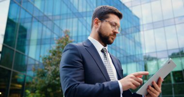 Tablet kullanırken sokakta yürüyen, resmi giyinmiş, arka planda modern bir binası olan beyaz bir adamın yan görüntüsü..