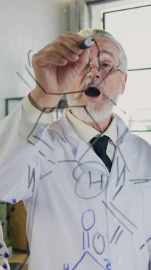 Çekici, saygı duyulan, sakallı, kimyasal formülün yapısını açıklayan, genç erkek ve kadın meslektaşlarına işaretleyici kullanarak çizdiği, klinikteki ortak toplantı sırasında.