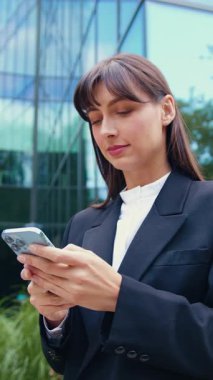 Siyah ceketli gülümseyen esmer kadın akıllı telefon kullanıyor ve açık havada güneşli bir yaz gününde modern cam bir binanın önünde yürürken mesaj yazıyor.