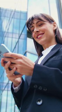 Kendine güvenen esmer, siyah ceketli bir iş kadını akıllı telefon kullanırken ve modern cam ofis binasının önünde mesaj yazarken dışarıda dolaşıyor.