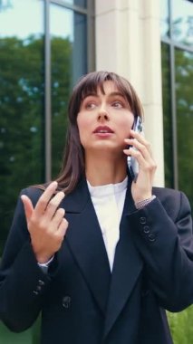 Siyah ceketli, esmer, beyaz bir kadın modern cam ofis binasının önünde duruyor. Teneffüste telefonla konuşuyor. Uzaktan kumandalı, açık havada çalışıyor.