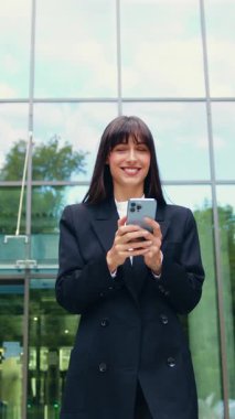 Resmi takım elbiseli, kendine güvenen genç ve gülümseyen bir iş kadını, iş çıkışı ve açık havada akıllı telefonunu kullanırken cam ofis binasından güvenle çıkıyor.