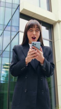 Şaşırtıcı iş kadınının elinde akıllı telefon var, bildirim aldığı için mutlu, ofisin arka planına karşı mutluluktan hareketler yapıyor, dışarıda. Duygular ve hoş duygular konsepti