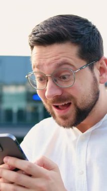 Beyaz tişörtlü çekici bir genç elinde telefonla başarıyı kutluyor, cep telefonu ekranına bakıyor, modern cam bina arka planında heyecanla yumruğunu kaldırıyor.