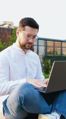 Modern cam bina arka planında çalışan serbest çalışan genç bir erkek. Çekici adam dışarıda dizüstü bilgisayarında sosyal medya sohbeti yapıyor.