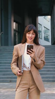 Şehirde bir iş gününden sonra modern bir ofis binasının önünde dikilirken resmi kıyafetli gülümseyen profesyonel bir kadın akıllı telefon kullanıyor.