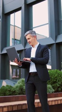 Çevrimiçi iş için bilgisayarla uzaktan kumanda kullanan yakışıklı yaşlı girişimci. Çalışan, kaydıran, dizüstü bilgisayarda çalışan, dışarıda ofis binasında oturan olgun, başarılı bir iş adamı.