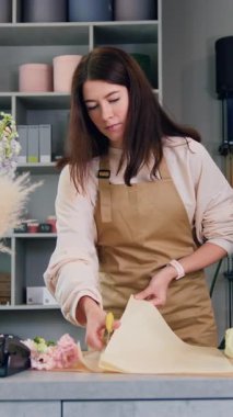 Önlüklü çiçekçi kadın zarif buket yapar. Çiçekçi kadın çiçek toplar, çiçekçide çalışan ambalaj kağıtlarını keser..