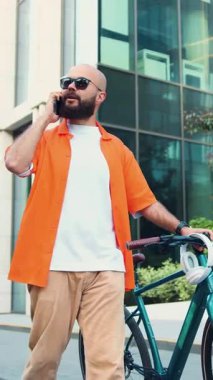 Yakışıklı, beyaz sakallı, sıradan kıyafetleri olan bir adam büyük şehir binasının yanında işe giderken bisikletle şehirde telefonla konuşuyor. Yavaş çekim.