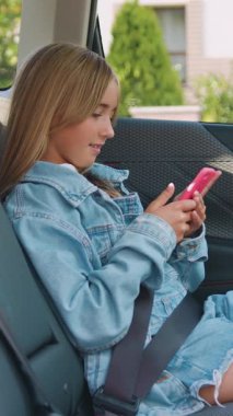 Şirin, mutlu, mutlu, 12 yaşında, şık kotlu, arka koltukta oturan, emniyet kemerini takan ve telefonda arkadaşlarıyla sohbet eden güzel bir kız.