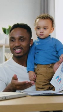 Video konferansı sırasında küçük çocuğuyla birlikte bilgisayarın önünde oturan yakışıklı, pozitif, yetenekli, Afrikalı Amerikalı işçiyle uzak çalışma.