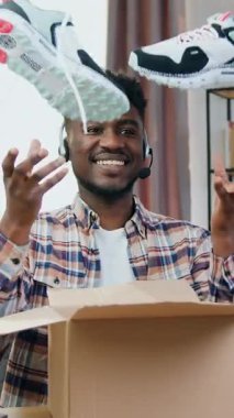 Yakışıklı, gülümseyen, siyah tenli, kablosuz kulaklıklı adam internet izleyicileri için video kaydederken teslimat paketindeki yeni spor ayakkabılarını gösteriyor.