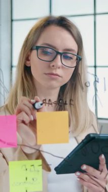 Modern ofisin cam duvarında çalışan güzel, yaratıcı sarışın kadın işçi bu duvara tablet cihazından bilgi kaydediyor ve en iyi finansal sonuçları nasıl elde edeceğimizi düşünüyor.